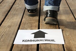 Schild Kommunalwahl mit Pfeil, im Hintergrund sind Schuhe zu sehen.