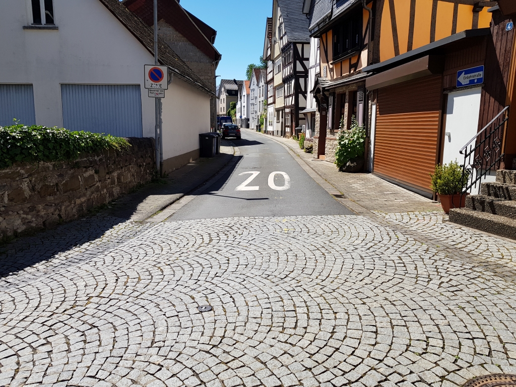 Bild der Chaldäergasse Herborn. Im Vordergrund ist die gepflasterte Einfahrt der Gasse zu sehen, im Hintergrund die Oberfläche aus Asphalt, die durch Pflastersteine ersetzt wird. 